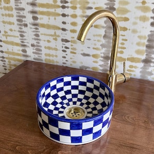 Puede incluir: Un lavabo redondo de cerámica a cuadros azules y blancos, con un grifo dorado. El lavabo tiene un desagüe de latón y está sobre una superficie de madera. El grifo es curvo. El fondo presenta un papel pintado con dibujos.