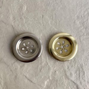 Two decorative, circular metal drain covers. One is silver-toned, the other is gold-toned. Each has a floral-shaped design in the center with small, curved cutouts. The drain covers are designed to fit into a sink or tub.