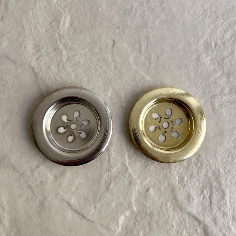 Two decorative, circular metal drain covers. One is silver-toned, the other is gold-toned. Each has a floral-shaped design in the center with small, curved cutouts. The drain covers are designed to fit into a sink or tub.