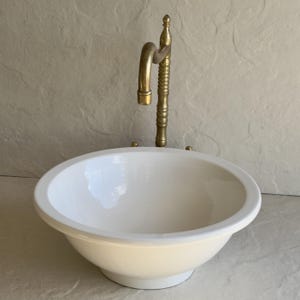 May include: A white, oval ceramic sink with a brass tap. The tap has a curved spout and a decorative, fluted column. The sink is set against a neutral background, suggesting a bathroom or washroom setting.