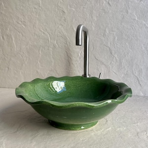 Puede incluir: Un lavabo de cerámica verde con un borde ondulado y un grifo de acero inoxidable. El lavabo tiene un acabado brillante y se asienta sobre una base de pedestal. El grifo es de diseño curvo con un solo mango.