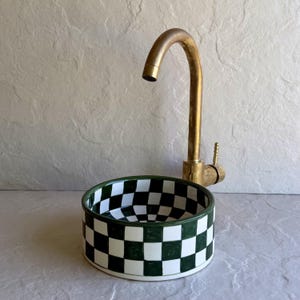 Peut inclure: Un évier en céramique avec un motif à carreaux verts et blancs. L'évier est associé à un robinet en laiton. Le robinet a un bec courbé et une seule poignée. L'évier est rond et a un large bord.