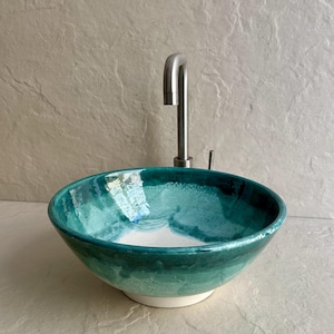 handcrafted ceramic vessel sink with a teal and white glaze. The round sink is paired with a brushed nickel faucet. The sink's interior features a blend of teal hues, creating a unique, artistic look for a bathroom.