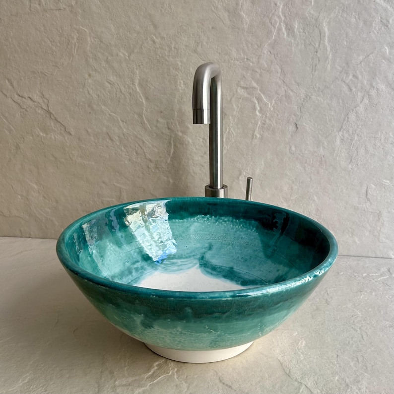 handcrafted ceramic vessel sink with a teal and white glaze. The round sink is paired with a brushed nickel faucet. The sink's interior features a blend of teal hues, creating a unique, artistic look for a bathroom.