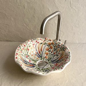 Könnte beinhalten: Ein weißes Keramikwaschbecken mit floralem Muster und einem Wasserhahn aus Edelstahl. Das Waschbecken hat einen gewellten Rand und ist mit bunten Blumen und Blättern in Rot-, Orange-, Lila- und Grüntönen verziert. Der Wasserhahn hat ein gebogenes Design.