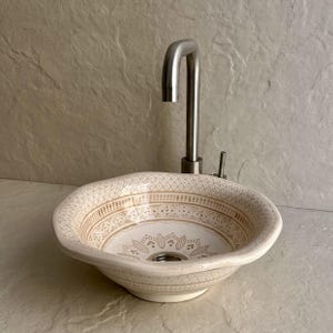 May include: A cream-coloured ceramic sink with a detailed brown floral pattern. The sink is paired with a brushed nickel tap. The sink's design features intricate geometric and floral motifs, adding a touch of elegance to any bathroom.