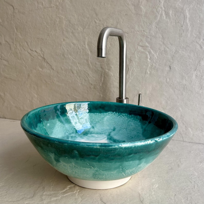 teal ceramic vessel sink with a glossy finish. The sink has a rounded bowl shape and a white base. A stainless steel faucet is mounted above the sink, with a curved spout.