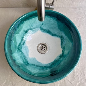 round ceramic sink with a teal and white glaze. The sink has a brushed metal drain and is mounted under a brushed metal faucet. The interior of the sink is white, with teal and turquoise brushstrokes around the edges.