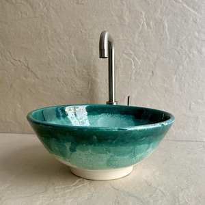 Puede incluir: Un lavabo de cerámica verde y turquesa con un grifo de acero inoxidable. El lavabo tiene forma de cuenco redondeado con una base blanca. El color pasa de un verde turquesa intenso a un verde más claro. El grifo es alto y curvado.