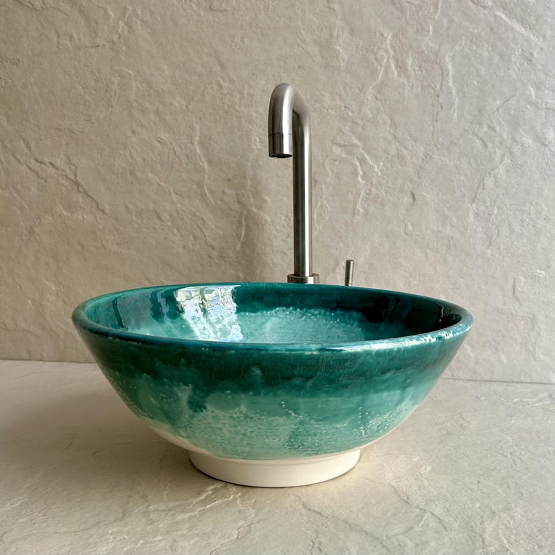 teal and green ceramic vessel sink with a stainless steel faucet. The sink has a rounded bowl shape with a white base. The color transitions from a deep teal to a lighter green. The faucet is tall and curved.