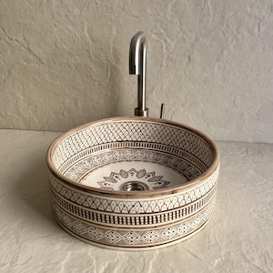 Peut inclure: Évier rond en céramique avec un motif décoratif marron et blanc. L'évier est équipé d'un robinet et d'un drain en acier inoxydable. Le design de l'évier présente des motifs géométriques et floraux, adapté à une salle de bain.