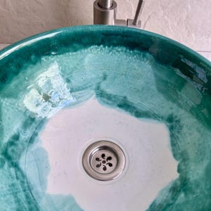 handcrafted ceramic sink with a unique teal and white glaze. The sink features a circular design with a stainless steel drain and faucet. The teal glaze creates a marbled effect, adding a touch of artistic flair to any bathroom.