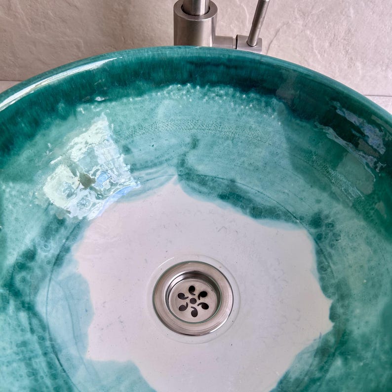 handcrafted ceramic sink with a unique teal and white glaze. The sink features a circular design with a stainless steel drain and faucet. The teal glaze creates a marbled effect, adding a touch of artistic flair to any bathroom.