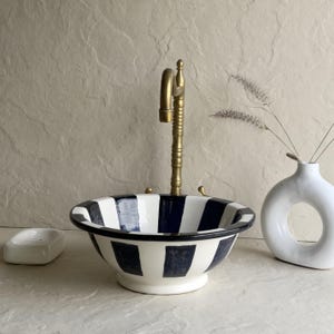 Puede incluir: Un lavabo de cerámica blanca con un diseño de rayas azul marino y blanco. Cuenta con un grifo de latón y está acompañado de un jabonera blanca y un jarrón blanco con una abertura circular. Un elemento decorativo para el baño.