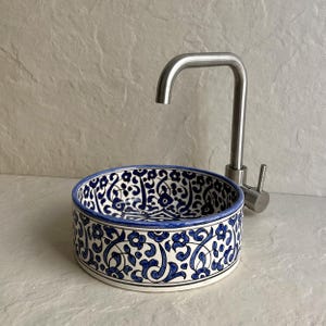 Pode incluir: Uma pia redonda de cerâmica com um padrão floral azul e branco. Uma torneira de aço inoxidável está posicionada acima da pia. A pia tem aproximadamente 30 cm de diâmetro.