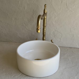 May include: A white, round ceramic sink with a brass faucet. The faucet has a curved spout and a decorative, ribbed column. The sink is a simple, modern design, suitable for a bathroom or powder room.