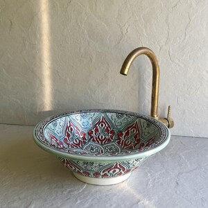 Puede incluir: Un lavabo de cerámica decorativo con un detallado diseño floral rojo, azul y blanco. El lavabo redondo tiene un borde verde y está junto a un grifo de latón. El lavabo está diseñado para un baño.