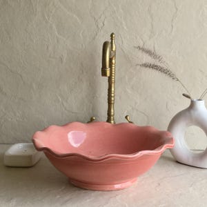 Peut inclure: Un évier en céramique rose avec un bord festonné et un robinet en laiton. L'évier est d'un rose doux et a une forme ronde. Un porte-savon blanc et un vase décoratif sont également visibles.