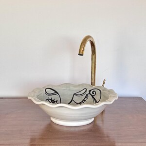 Peut inclure: Un lavabo en céramique gris clair avec un bord festonné et un motif de visage noir. Le lavabo est doté d'une base blanche et d'un robinet doré. Idéal pour une salle de bain.