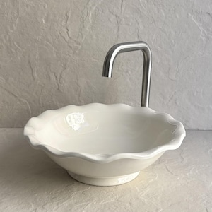 Può includere: Un lavabo in ceramica bianca con bordo smerlato e rubinetto in acciaio inossidabile. Il lavabo ha una forma rotonda a forma di ciotola e poggia su un piccolo piedistallo. Il rubinetto ha un beccuccio curvo.