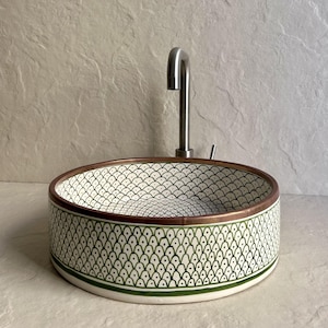 Può includere: Un lavabo rotondo in ceramica con un motivo decorativo di forme a goccia verdi e bianche. Il bordo ha una fascia color rame. Un rubinetto in acciaio inossidabile è installato sopra il lavabo.