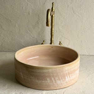 May include: A peach-coloured ceramic sink with a gold-toned rim and a matching tap. The sink has a simple, modern design. The tap has a curved spout and two small handles. The background is a light beige stone.