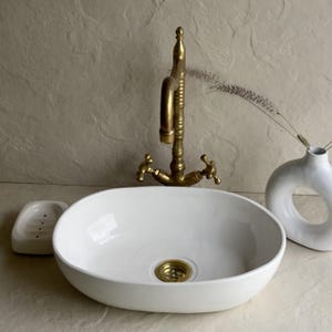 May include: A white oval ceramic sink with a gold-colored drain and faucet. The faucet has cross handles and a curved spout. A white soap dish and a white vase with a circular opening are also present.
