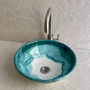 round ceramic vessel sink with a vibrant teal and white glaze. The sink features a stainless steel faucet and drain. The interior has a white base with teal accents, and the rim is a darker teal. The sink is designed for bathroom use.