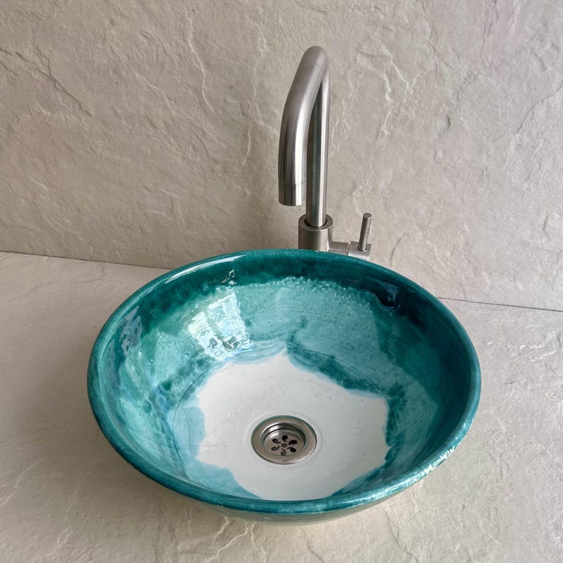 round ceramic vessel sink with a vibrant teal and white glaze. The sink features a stainless steel faucet and drain. The interior has a white base with teal accents, and the rim is a darker teal. The sink is designed for bathroom use.