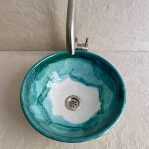 round ceramic sink with a vibrant teal and white glaze. The sink has a stainless steel drain and faucet. The color transitions from a deep teal at the rim to white in the center, creating a watercolor effect.