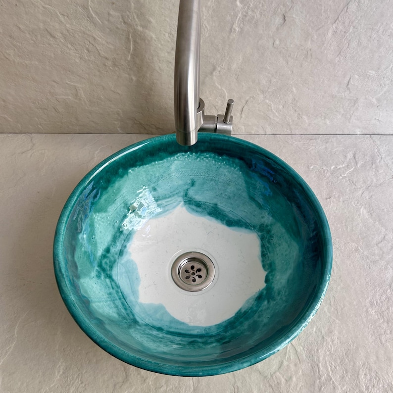 round ceramic sink with a vibrant teal and white glaze. The sink has a stainless steel drain and faucet. The color transitions from a deep teal at the rim to white in the center, creating a watercolor effect.