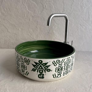 Peut inclure: Un évier rond en céramique blanche avec un intérieur vert et un robinet en acier inoxydable. L'extérieur de l'évier présente un motif géométrique vert foncé répétitif. Le robinet est de conception moderne et incurvée.