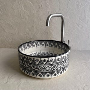Puede incluir: Un lavabo redondo de cerámica blanca con un patrón geométrico negro y un grifo de acero inoxidable. El borde del lavabo y los detalles del patrón son negros. El grifo tiene un caño curvo.