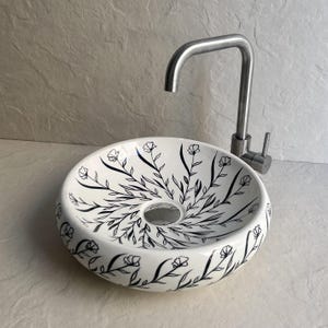 Peut inclure: Un lavabo en céramique blanche avec un motif floral noir et un robinet en acier inoxydable. L'évier rond présente un trou de vidange central. Le robinet a un bec courbé et une poignée à levier unique.