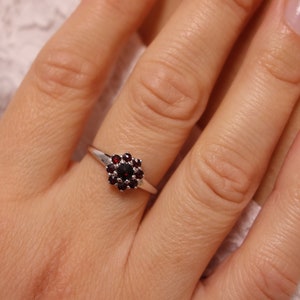 Puede incluir: Un anillo de plata con un grupo de pequeñas piedras preciosas rojas en el centro.
