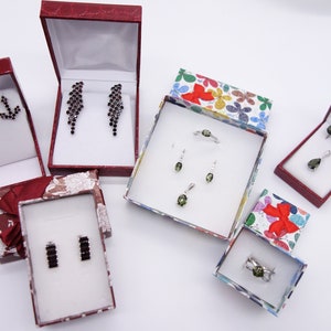 May include: Five small boxes with different colored lids, each containing a set of earrings or a ring. The boxes are on a white surface. The jewelry is made of silver and has green and black gemstones.