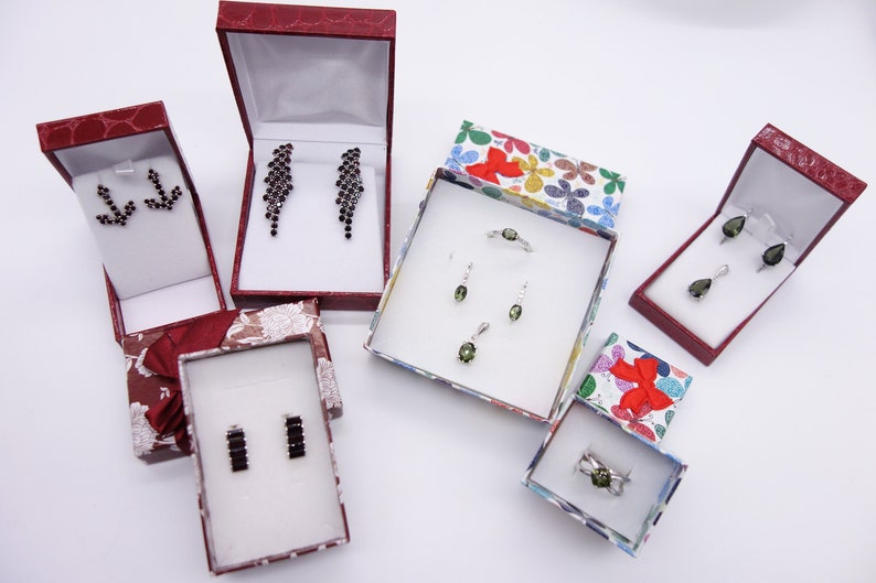 May include: Five small boxes with different colored lids, each containing a set of earrings or a ring. The boxes are on a white surface. The jewelry is made of silver and has green and black gemstones.