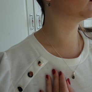 May include: A white sweater with gold buttons and a silver chain necklace with a green gemstone pendant. The person is wearing a ring on their finger and has red nail polish.