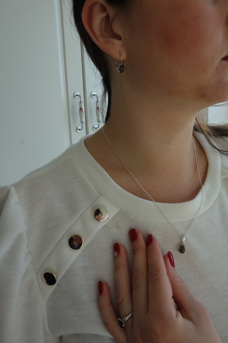 May include: A white sweater with gold buttons and a silver chain necklace with a green gemstone pendant. The person is wearing a ring on their finger and has red nail polish.