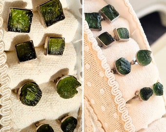May include: A close-up of two sets of earrings with green gemstone studs. The earrings are displayed on a white fabric background with a decorative border.