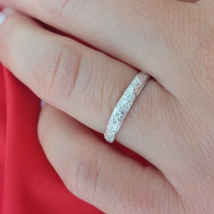 May include: A silver band ring with an intricate floral design. The ring is worn on a finger.