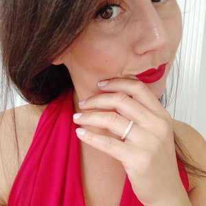 May include: A close-up of a woman's hand wearing a thin silver band ring with small sparkling stones. The woman is wearing a red halter top and has red lipstick on.