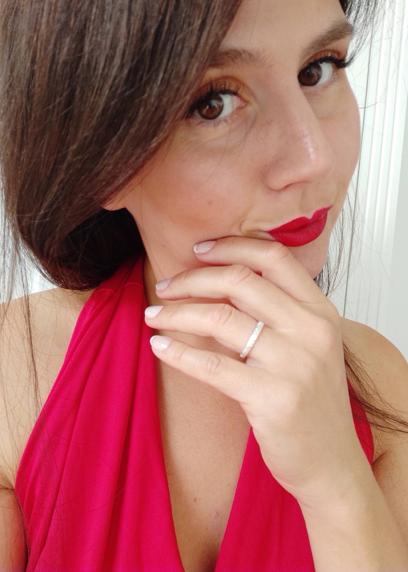 May include: A close-up of a woman's hand wearing a thin silver band ring with small sparkling stones. The woman is wearing a red halter top and has red lipstick on.