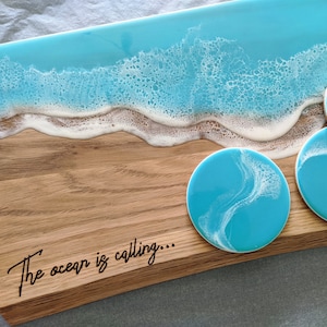 May include: Wooden serving board and matching coasters with a beach-themed design. The board features a blue resin ocean design with white foamy waves. The coasters have a similar blue and white swirl pattern. The text "The ocean is calling..." is on the board.