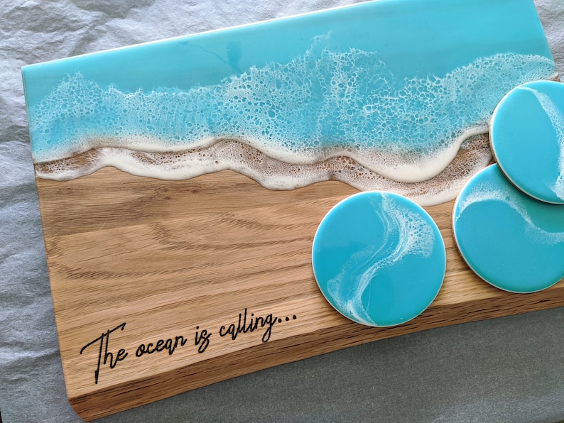 May include: Wooden serving board and matching coasters with a beach-themed design. The board features a blue resin ocean design with white foamy waves. The coasters have a similar blue and white swirl pattern. The text "The ocean is calling..." is on the board.