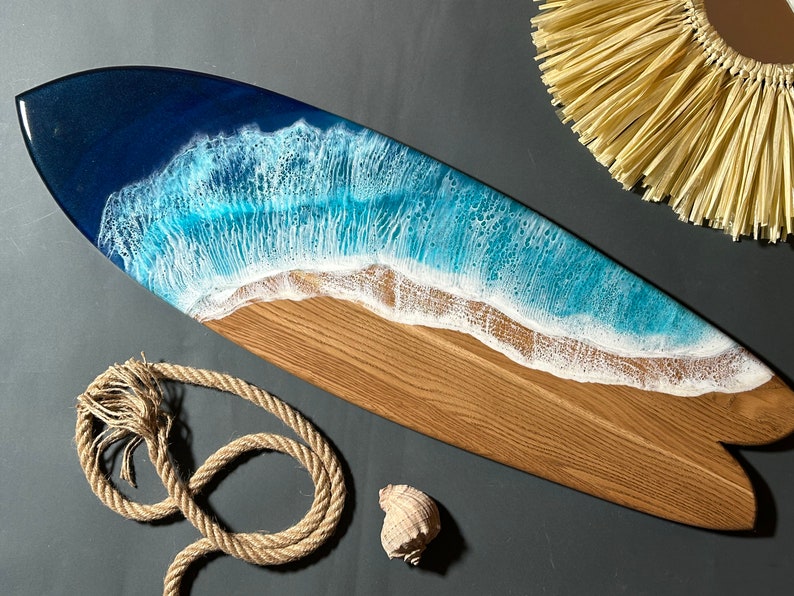 Op de afbeelding: Een houten surfboard met een blauw en wit hars ocean wave design. Het surfboard is een decoratief stuk en is niet bedoeld om te surfen.