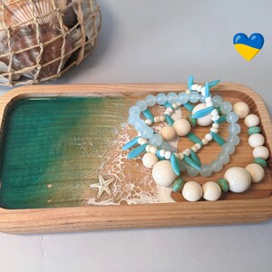 May include: A wooden tray with a green and white resin inlay depicting a beach scene. The tray features a starfish and three beaded bracelets in shades of blue and white.