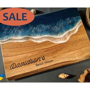 May include: Wooden cutting board with a beach-themed design. The top section features a blue and white wave pattern, transitioning to a light brown wood grain. The text "Davidson's Beach House" is inscribed on the board. An orange oval with the word "SALE" is in the top left corner.