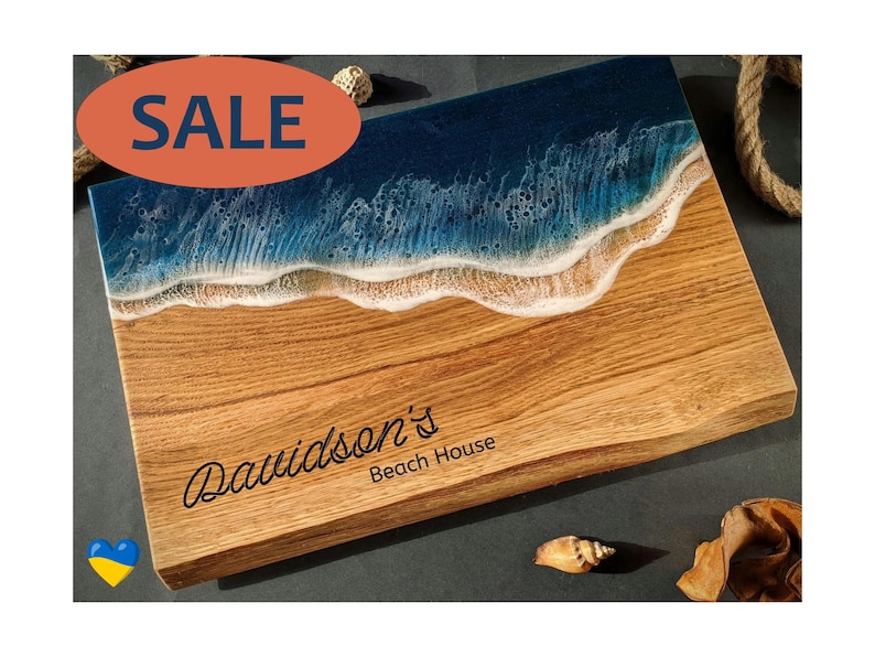May include: Wooden cutting board with a beach-themed design. The top section features a blue and white wave pattern, transitioning to a light brown wood grain. The text "Davidson's Beach House" is inscribed on the board. An orange oval with the word "SALE" is in the top left corner.