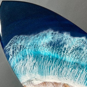 Op de afbeelding: Close-up van een kunstwerk van een surfboard van hars met een blauw en wit golfontwerp. De golf is gemaakt om eruit te zien als brekende oceaan golven met wit schuim.